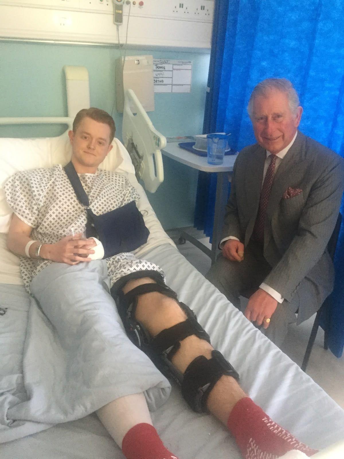 Travis Frain lays on a hospital bed with his leg strapped while King Charles is at his bedside. 