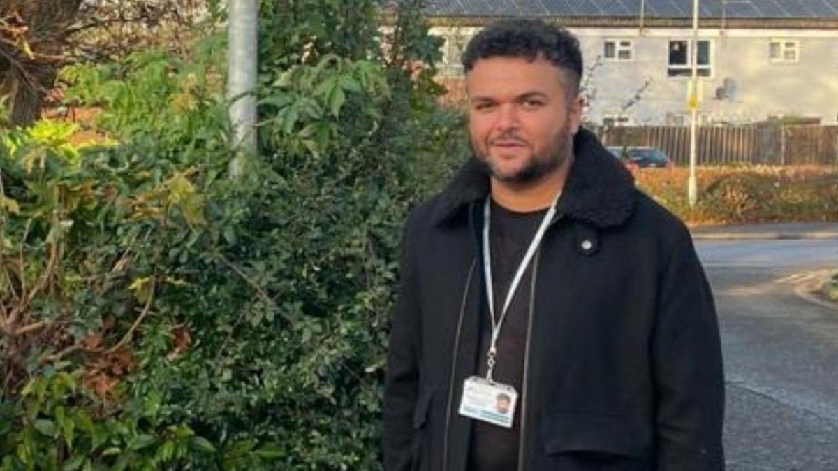 Alex Rafiq wearing a black blazer and green lanyard looking at the camera.