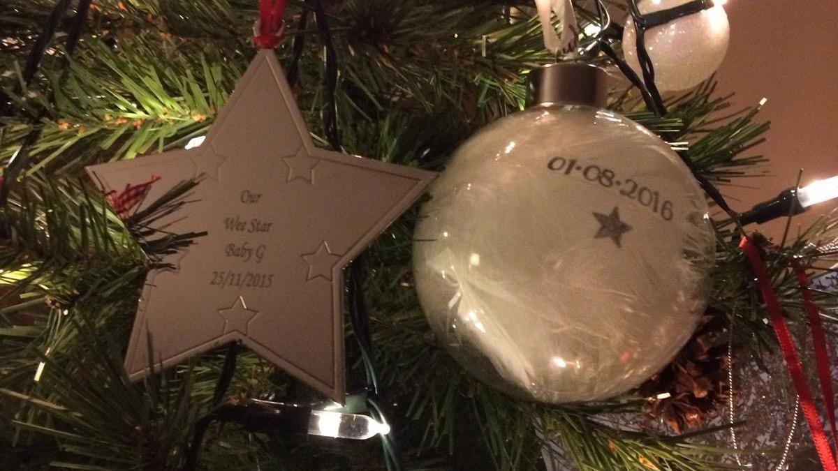 A close up image of a beige gold star and a Christmas bauble on a Christmas tree. Lights and some other decorations can be seen on the tree. 'Our wee star Baby G is written 25/11/2015' is inscribed on the star.