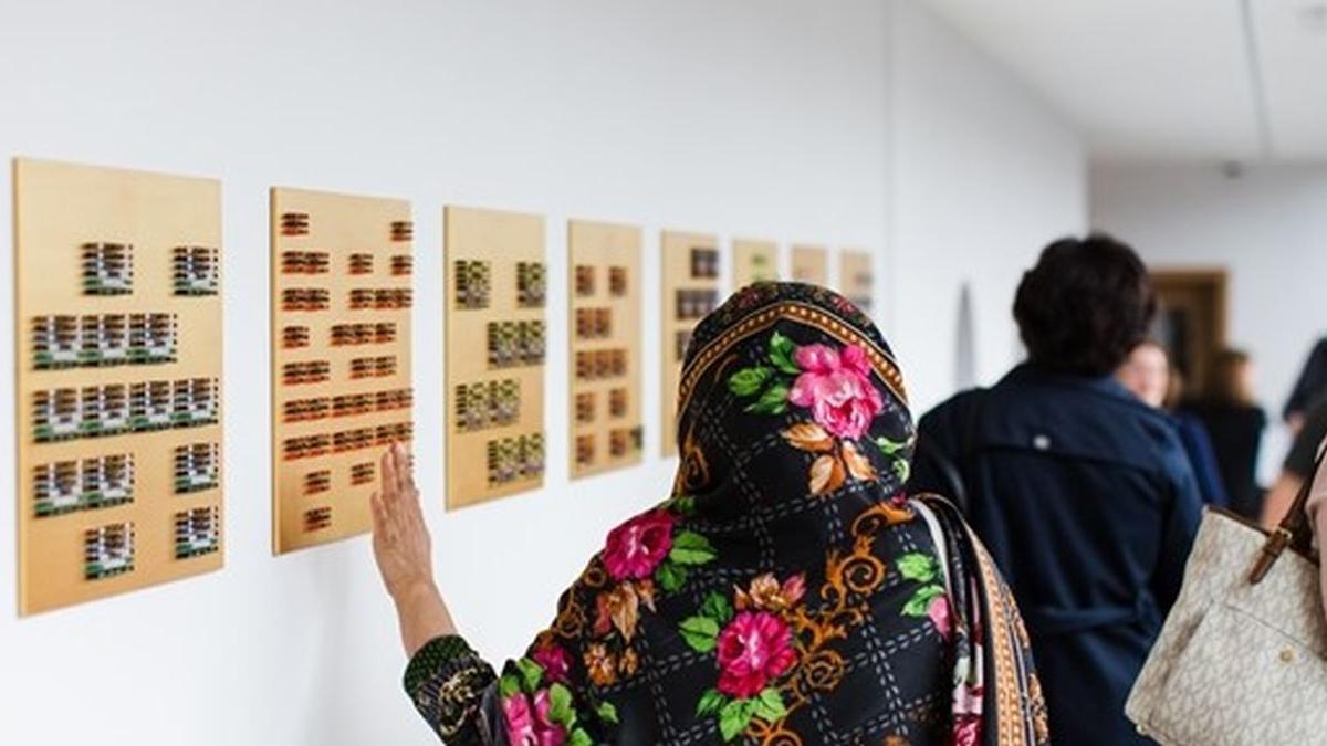 A woman in South Asian dress, looking at artwork in a gallery, her left arm is nearly touching the wall. She has her back to the camera, there are other people in the room but they are blurred or have their back to the camera. There are eight art images on a white wall. 