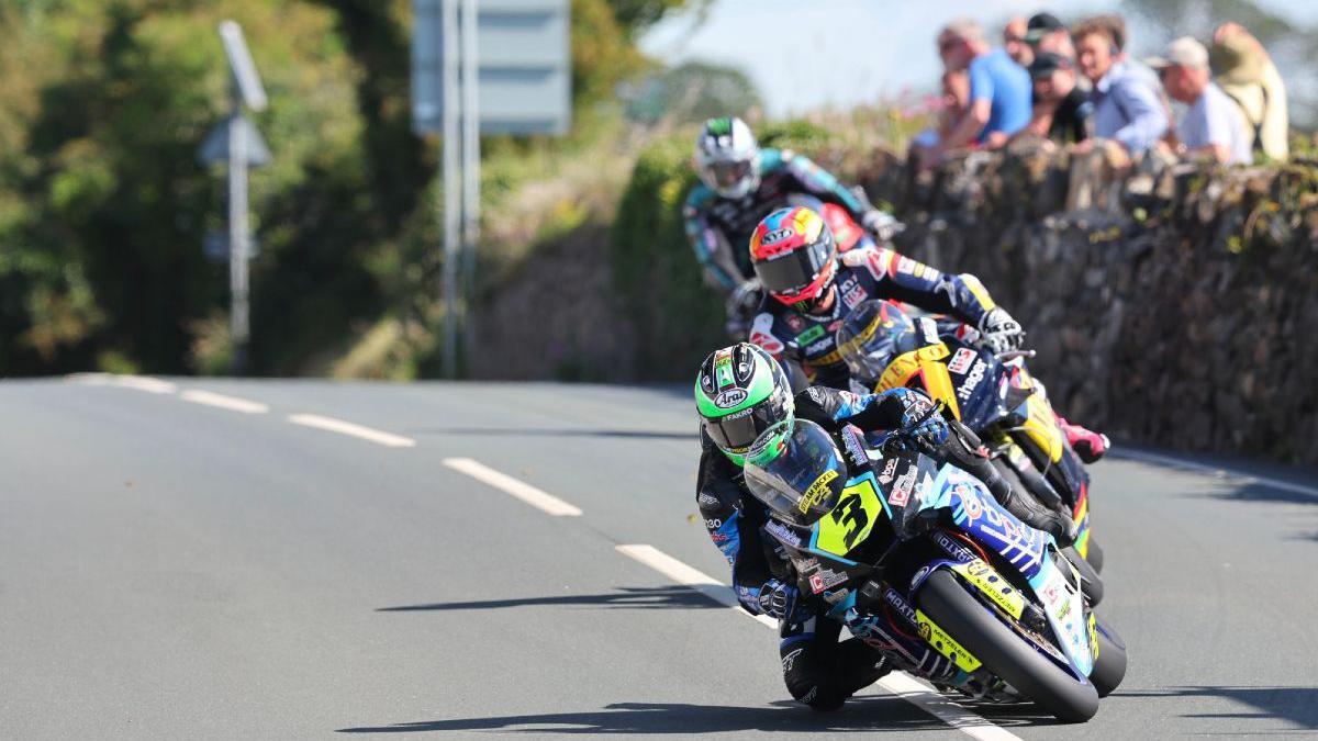 Mike Browne leading on the road with Davey Todd and Michael Dunlop behind  as they lean into a bend while spectators looks on from behind a stone wall.