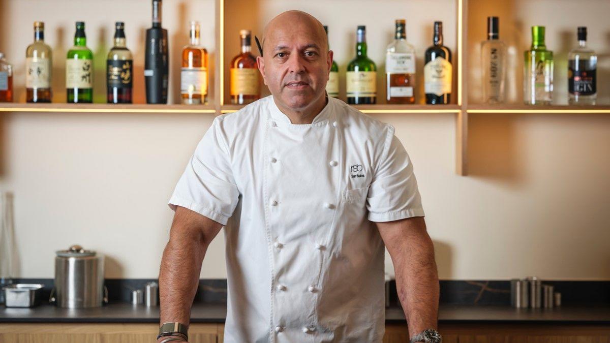 Sat Bains, wearing a chef's uniform, stands at a counter in his restaurant, with his hands on the surface. There is kitchen equipment behind him in silver containers and a line of bottles above him on a wall-mounted shelf.