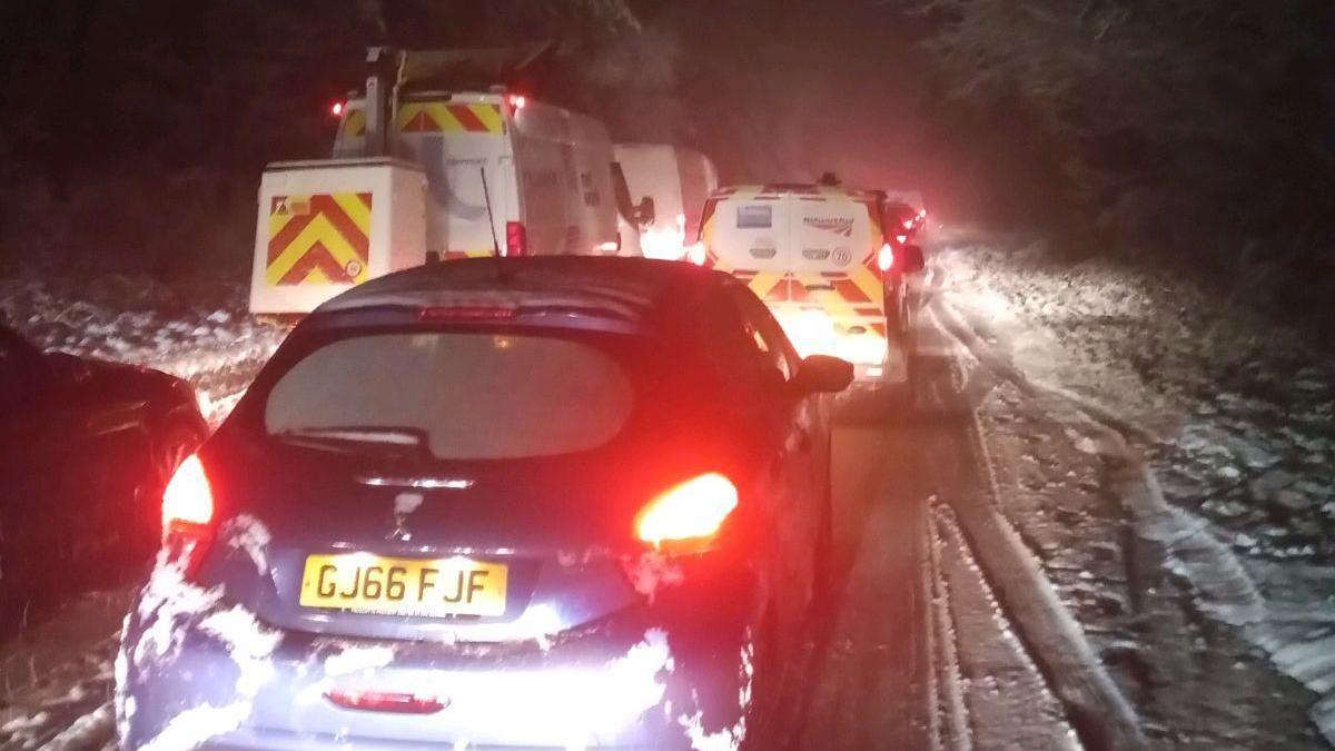 Multiple cars and vans are stuck on the A380 Telegraph Hill near Exeter after heavy snow fell in the area. It is dark and all the vehicles have their lights on.