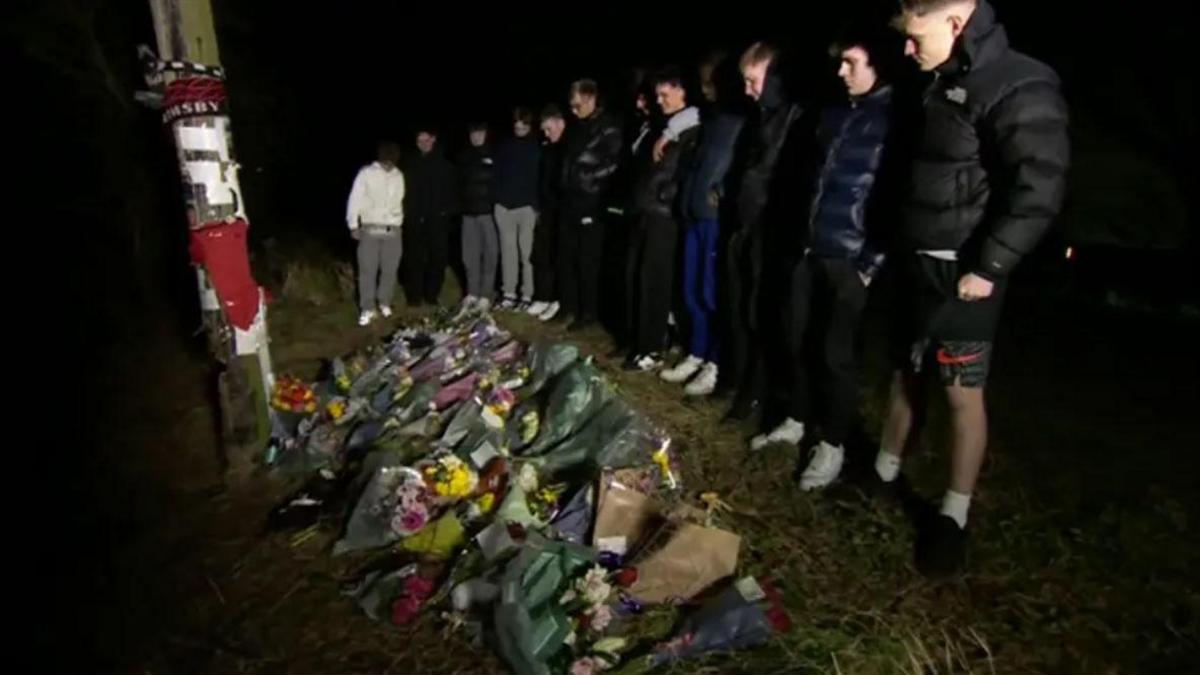 A group of lads wearing black coats and lined up looking down at floral tributes next to a post which has Grimsby football merchandise on it. It's night-time.