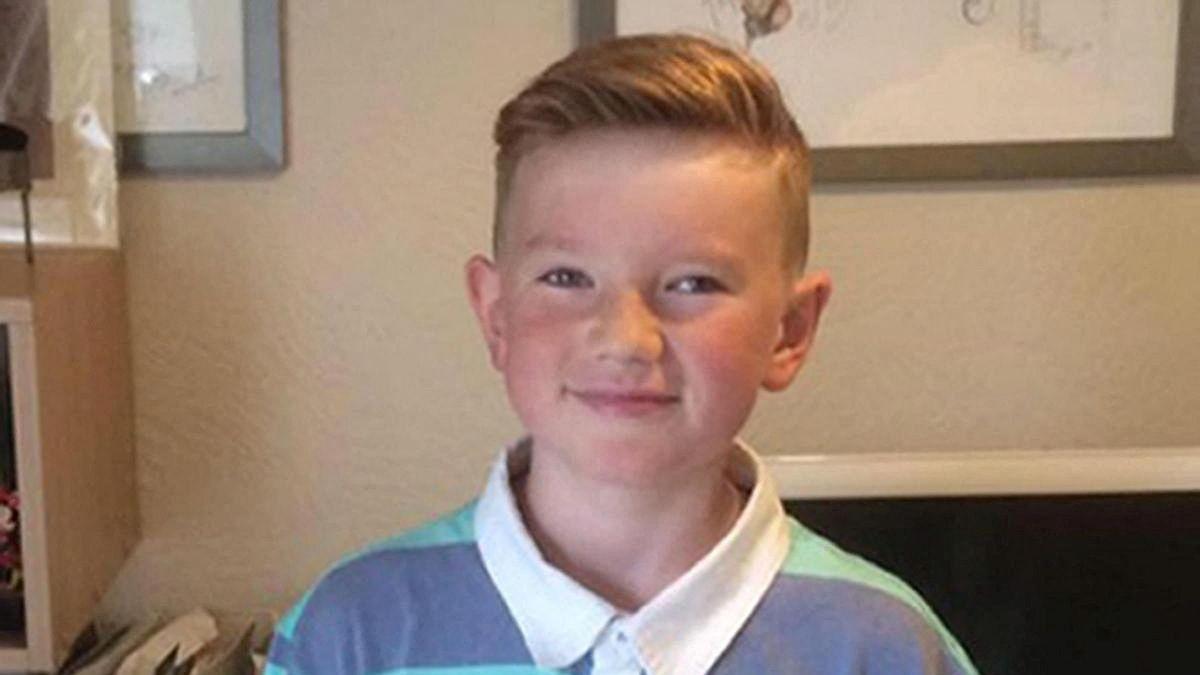 A picture of Alex Batty, age 11, wearing a white-collared polo shirt, smiling for a photograph in a living room. 