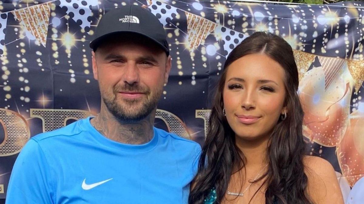 Tom Gaughan stands next to his daughter Alesha at a party. He is wearing a blue t-shirt and a black baseball cap. He has dark eyes and a beard. Alesha has long, dark brown hair and is smiling and wearing a sequin dress.