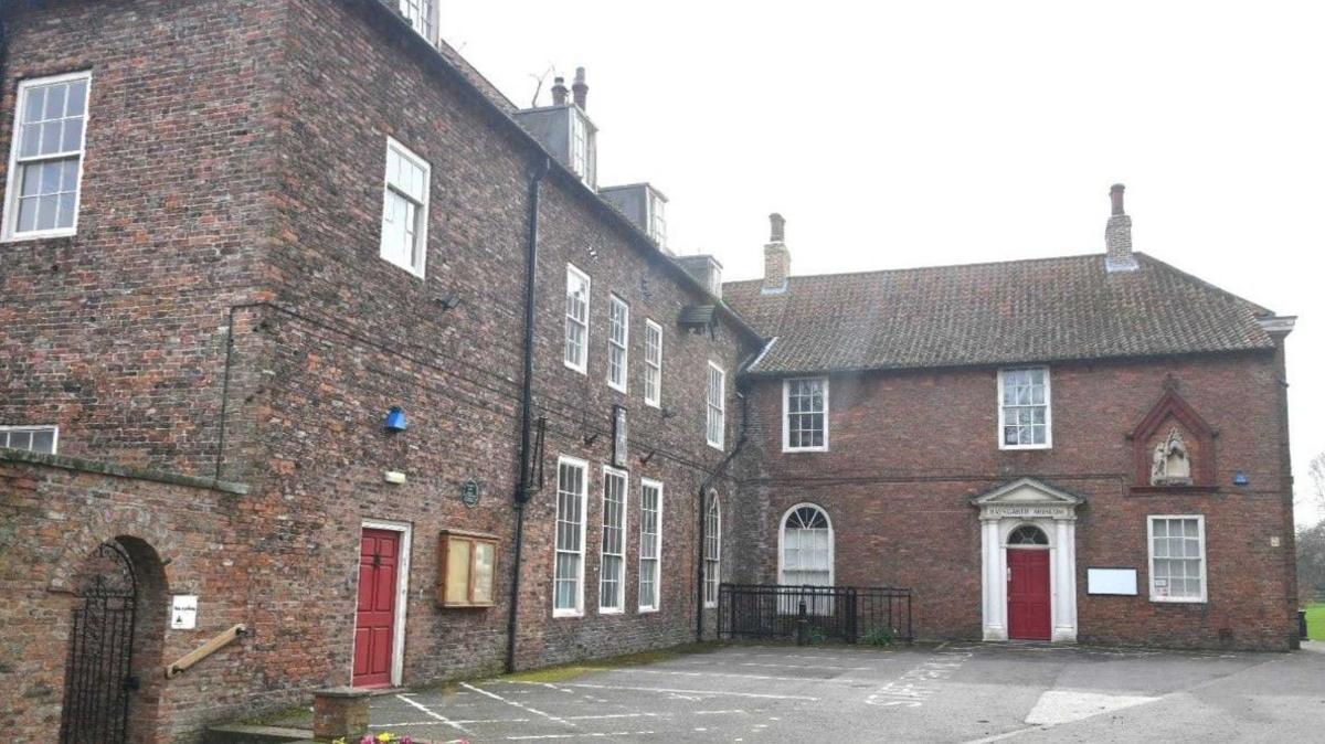 An image of the rear of the building, it has large old windows and more red painted doors. There is a tarmacked courtyard with parking space. 