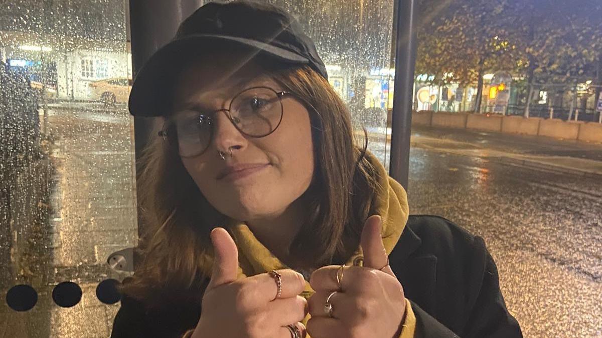 A young woman wearing glasses and a cap standing at a bus stop in the rain. It is dark, and the woman has her thumbs up.