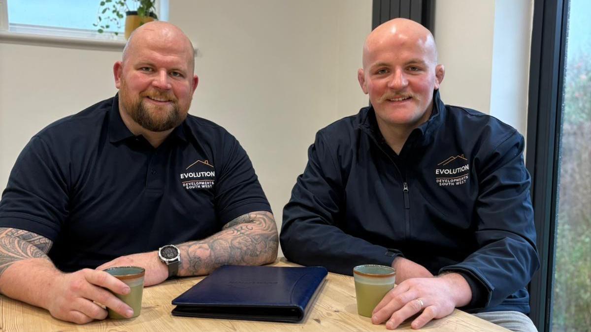 Jake, pictured on the left, sits next to his brother Josh at a wooden table. Jake wears a navy blue polo shirt with the Evolution Development South West logo on the right breast. He is holding a green coffee cup in one hand. Josh is on the right, also wearing a company jacket and holding a green coffee cup in his hand. They both smile at the camera.