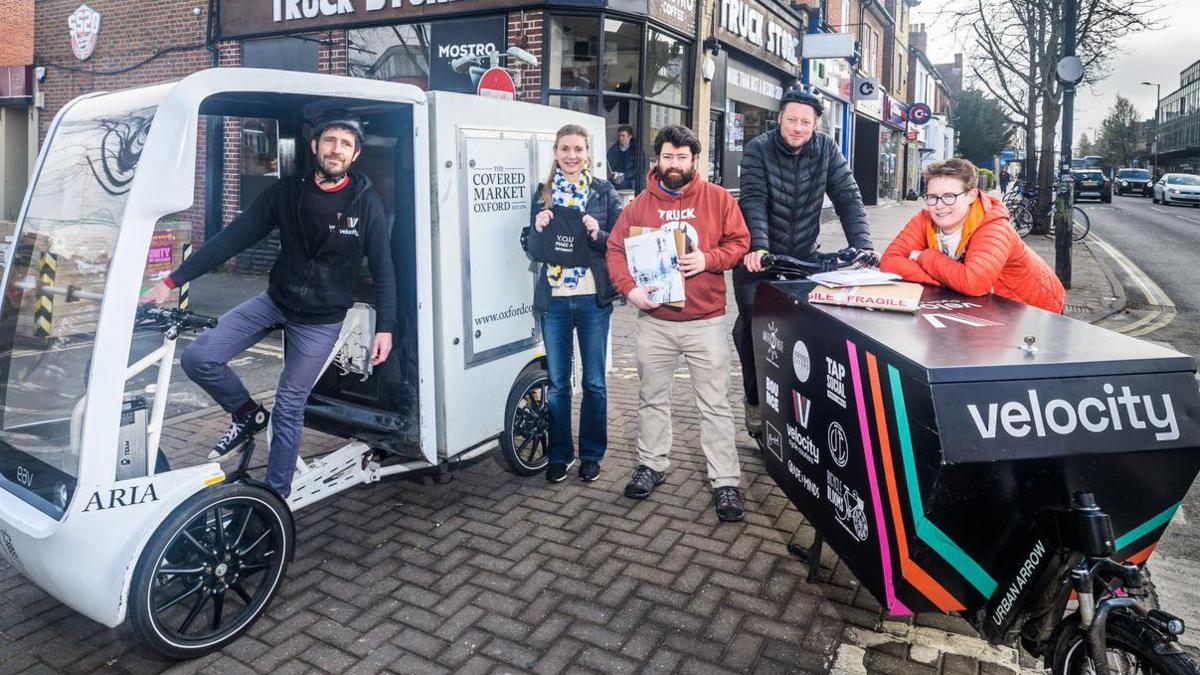 A man on a large white electric cargo bike is stood with four other people, including a man on a smaller black cargo bike.