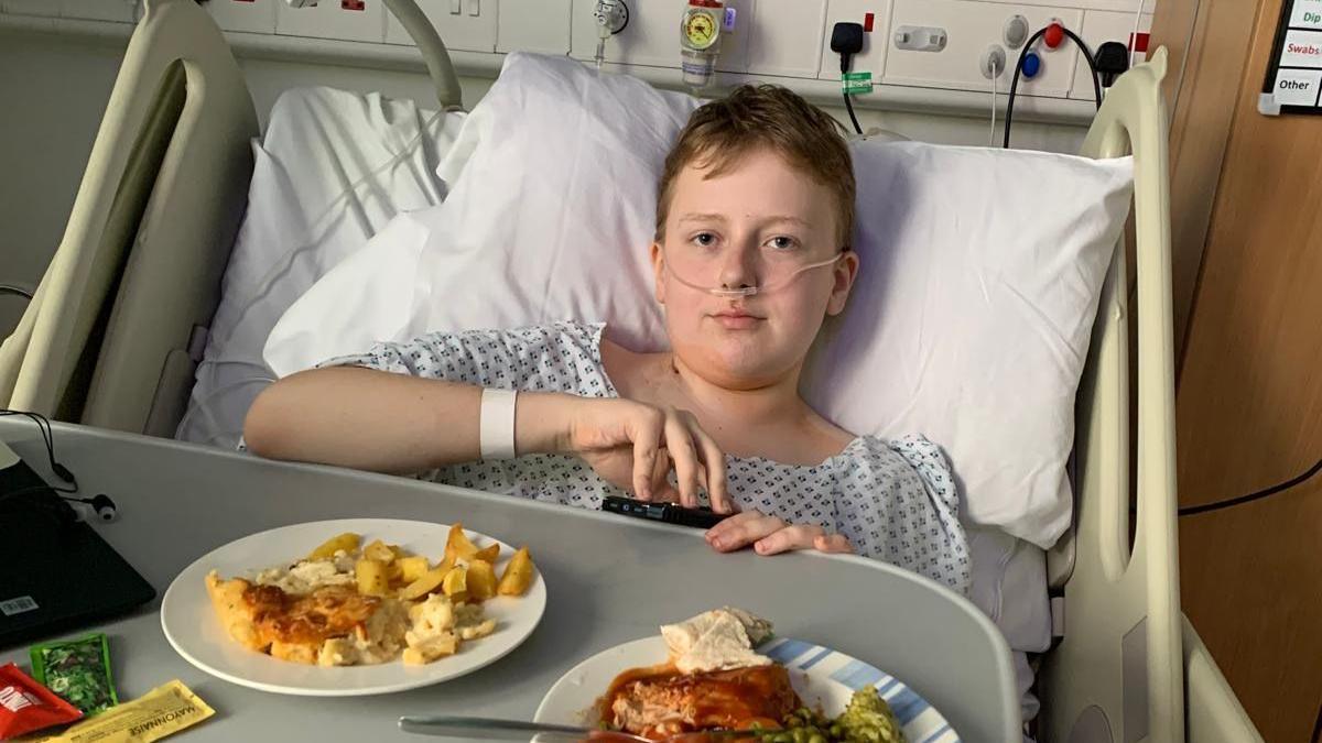 Adam is in a hospital bed wearing a hospital gow. He has an oxygen tube in his nose and the table has a plate of food in front of him.