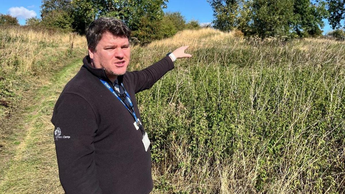 A man with short brown hair and wearing a black quarter zip jumper and a blue lanyard is stood in a field outside, surrounded by trees and bushes. 