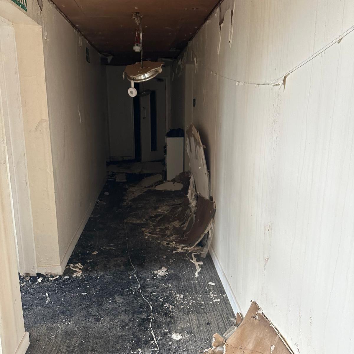 A fire-damaged corridor with debris and broken ceiling panels scattered on the floor, and a light fitting hanging loose from the ceiling.
