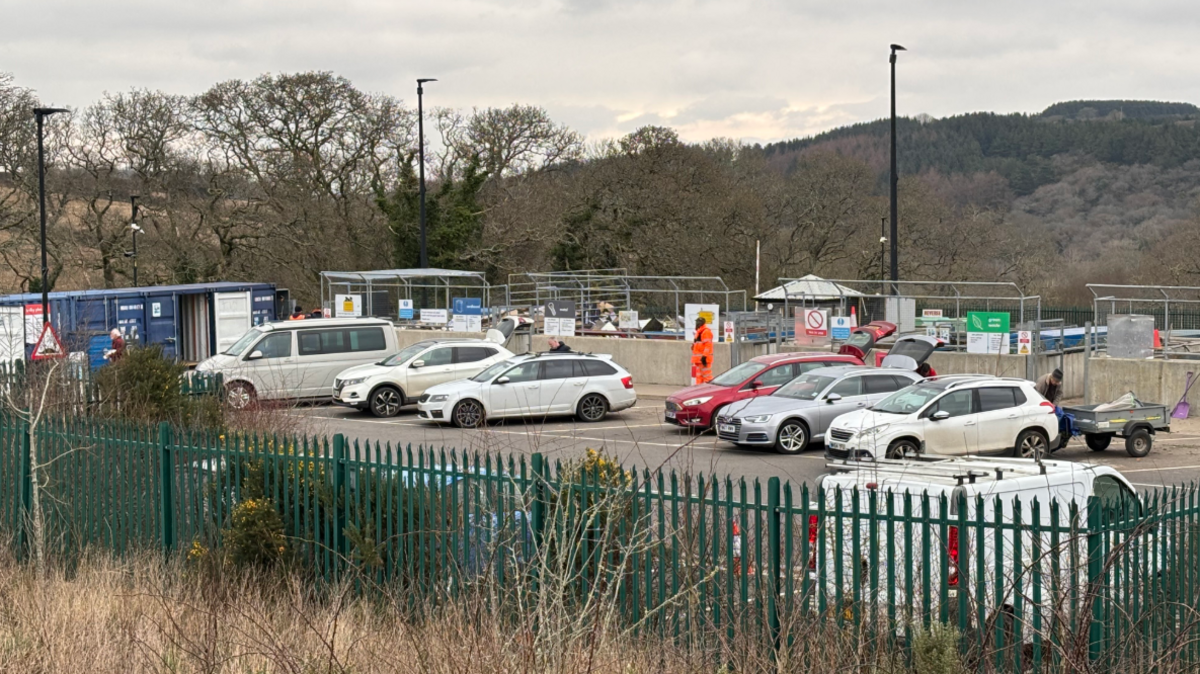 Truro's household waste recycling centre. The sky is grey and cloudy in the background and there are workers in the background. There are cars parked in the bays used by people bringing their recycling to the centre. 