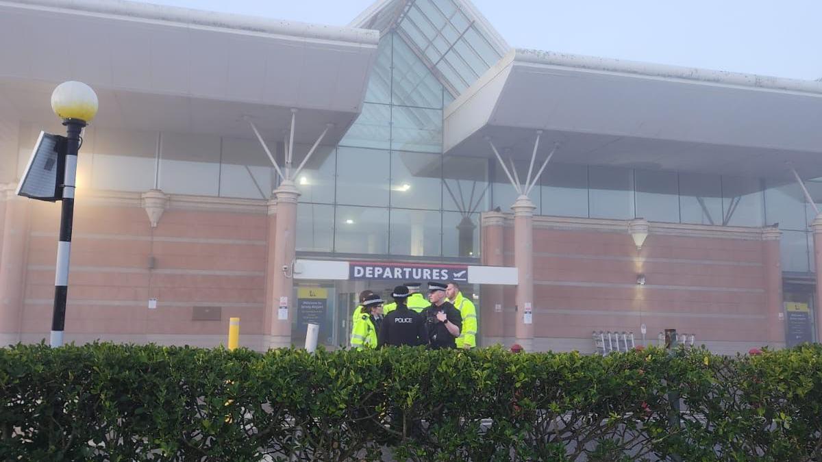 A group of police officers stand outside the entrance of Jersey Airport departures lounge. The weather is foggy. Some are wearing high vis jackets while others are dressed in black. 