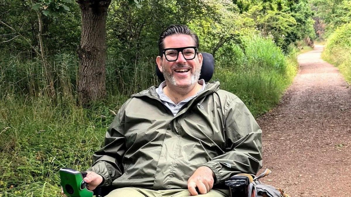 A man with dark hair and glasses wearing a green coat is sat in a wheelchair using a path that is lined by long grass and trees.