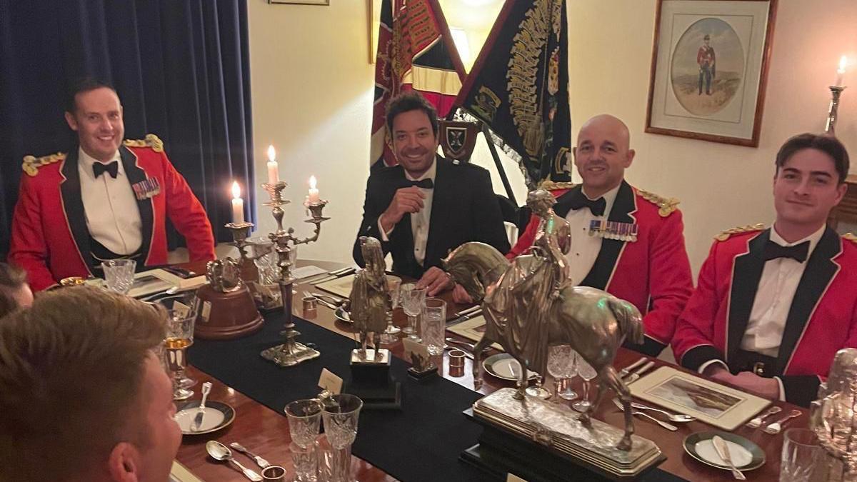 Five men sat around a dinner table with ornate decorations on it. Four of the men are wearing red blazers with bow ties and some are wearing medals.