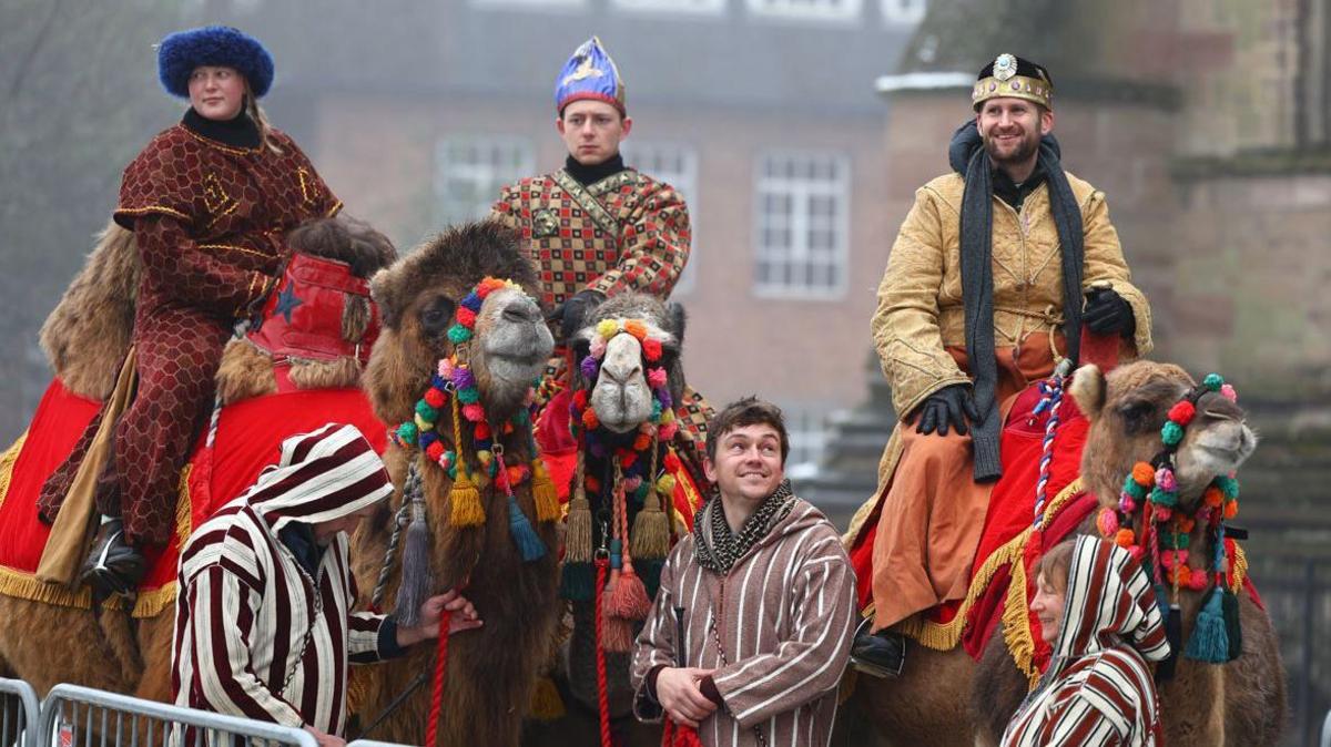 Three camels with red cloths covering their backs and colourful bridles being ridden by two men and a woman dressed as wise men, led by two men and a woman in striped robes. 