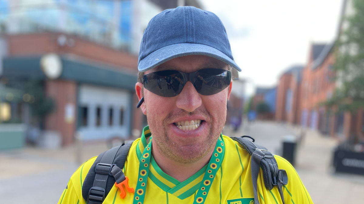 A head and shoulders image of Luke Ashman. He is grinning and wearing a Norwich shirt. He also has dark glasses and a hat on.