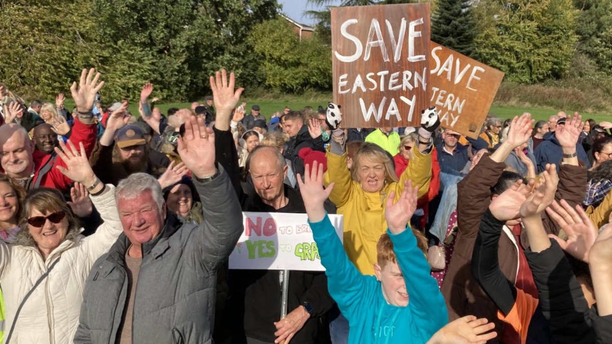 Dozens of men, women and children grouped together, some with raised hands, with a woman in a yellow coat holding a placard which reads Save Eastern Way. In the background can be seen grass and trees.