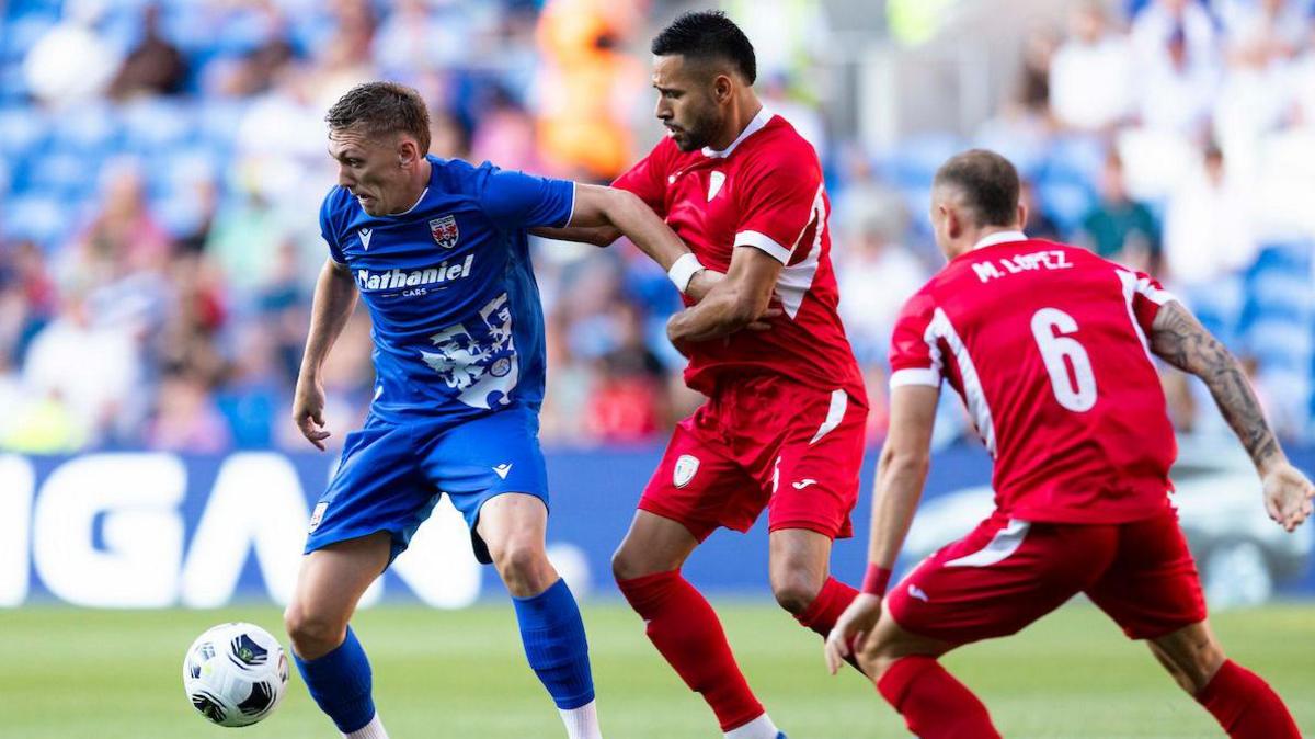 Nathan Wood in an all blue Penybont kit shields the ball from two Santa Coloma players in all red kit