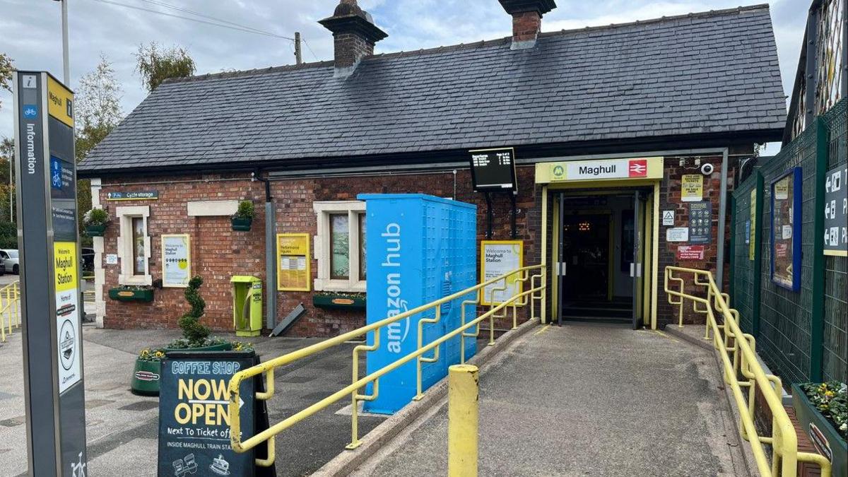 The entrance to Maghull railway station, with a sign for The Coffee Carriage coffee shop outside and hanging baskets with flowers on the wall. A planter with flowers can also be seen to the side of the ramped entrance.