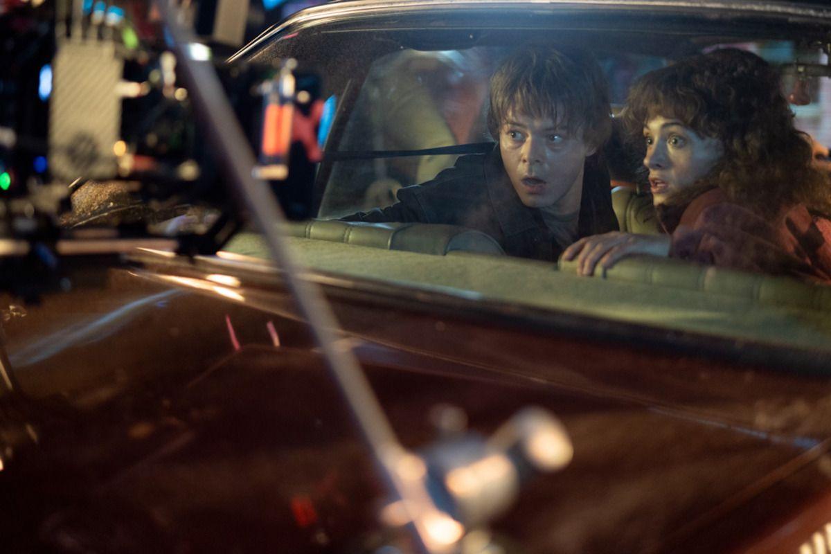 A behind-the-scenes shot of a young male and female actor in the back seat of a vintage car. They're looking out of the back window, with shocked expressions on their faces. In the foreground a camera rig capturing the pair's faces is just about visible.