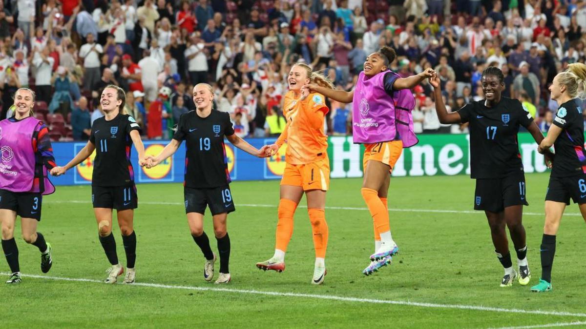 Six women in England kits hold hands while they run up a pitch in celebration. Crowds can be seen in the stands behind them.