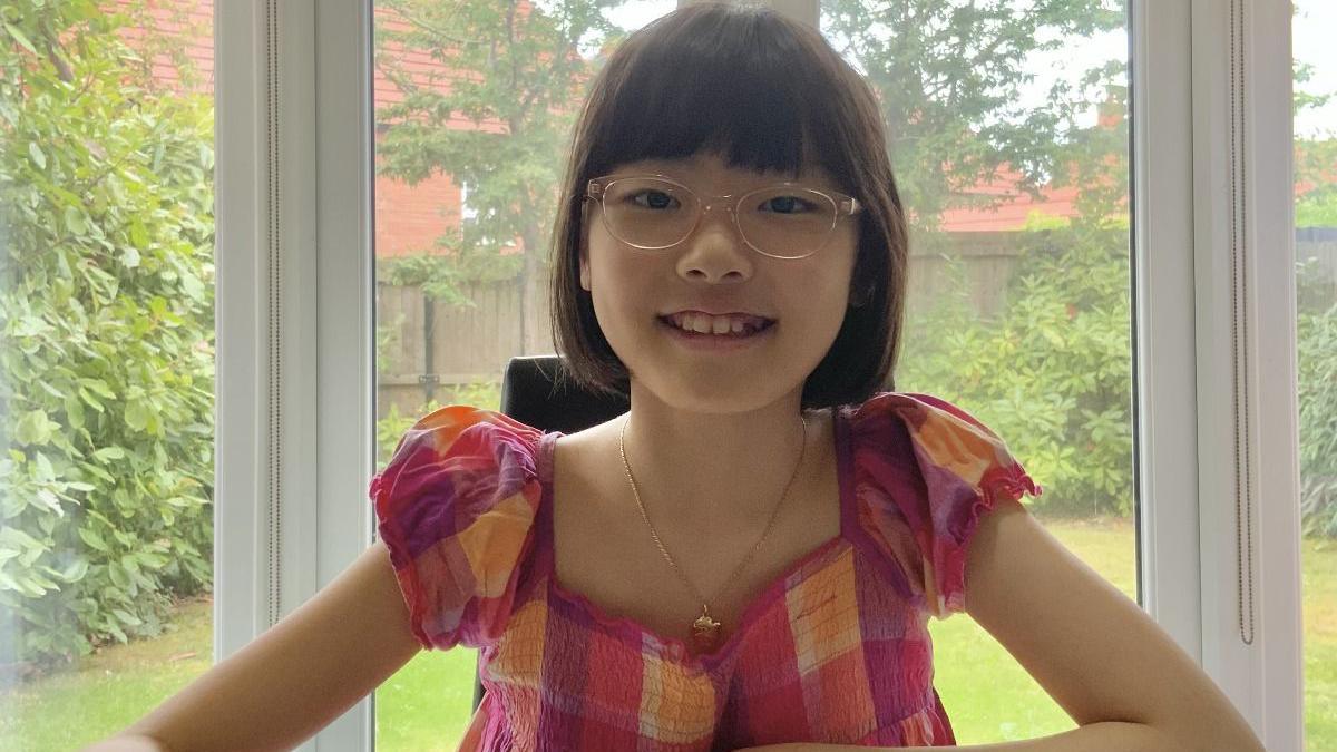 A girl with mid-length dark hair, glasses and a pink, purple and orange top, and wearing a gold necklace, smiles as she sits in a conservatory. A back garden can be seen through the window behind her.