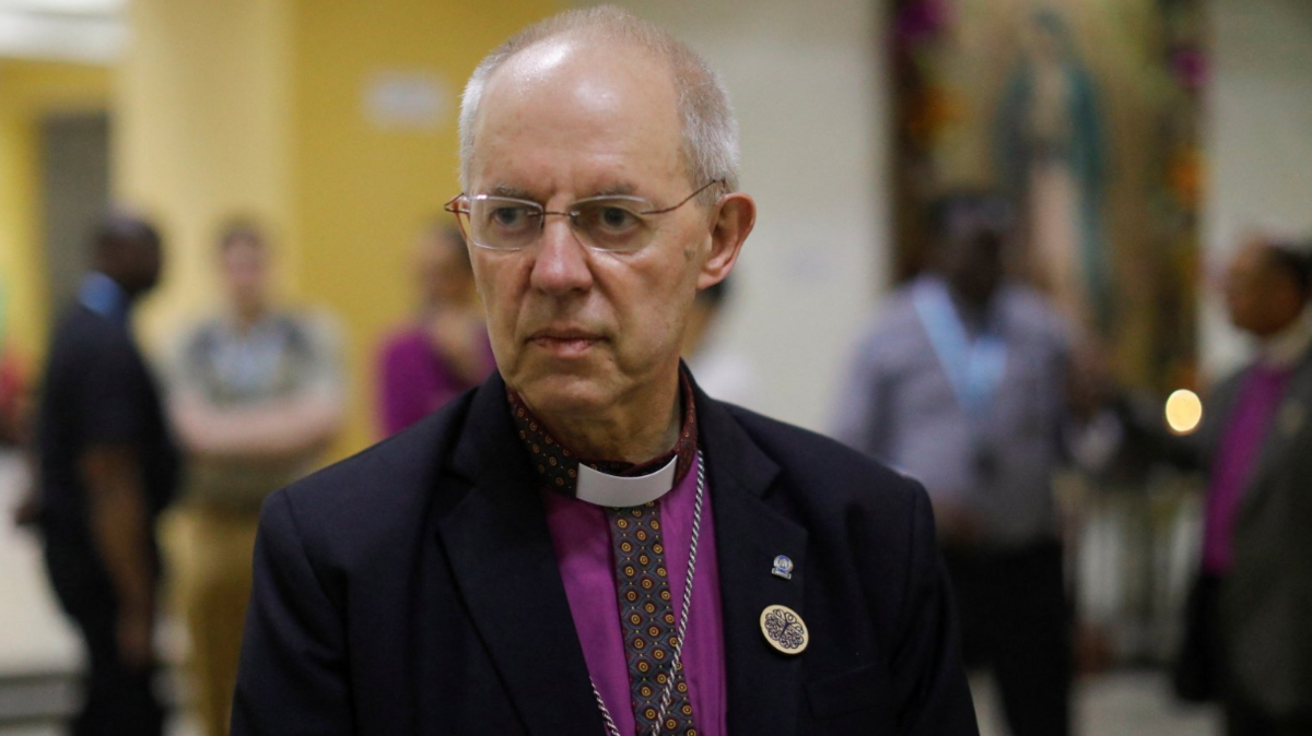 Archbishop of Canterbury Justin Welby looks on as he speaks with the press after a visit to the grave of Saint Oscar Arnulfo Romero, during a visit to El Salvador. Welby is wearing a black blazer with a bright purple shirt. A silver cross dangles from his neck against his chest.