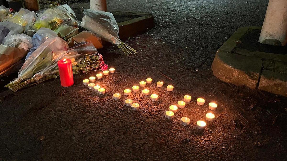 Candles spelling out "Leo" are left on the ground next to bunches of flowers on a dirt path next to a concrete bollard.