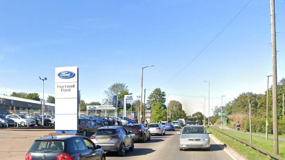 Redbourn Road, Hemel Hempstead, with cars on the road, by a car dealership. There are trees and grass to the left, and signs for the dealership to the right.
