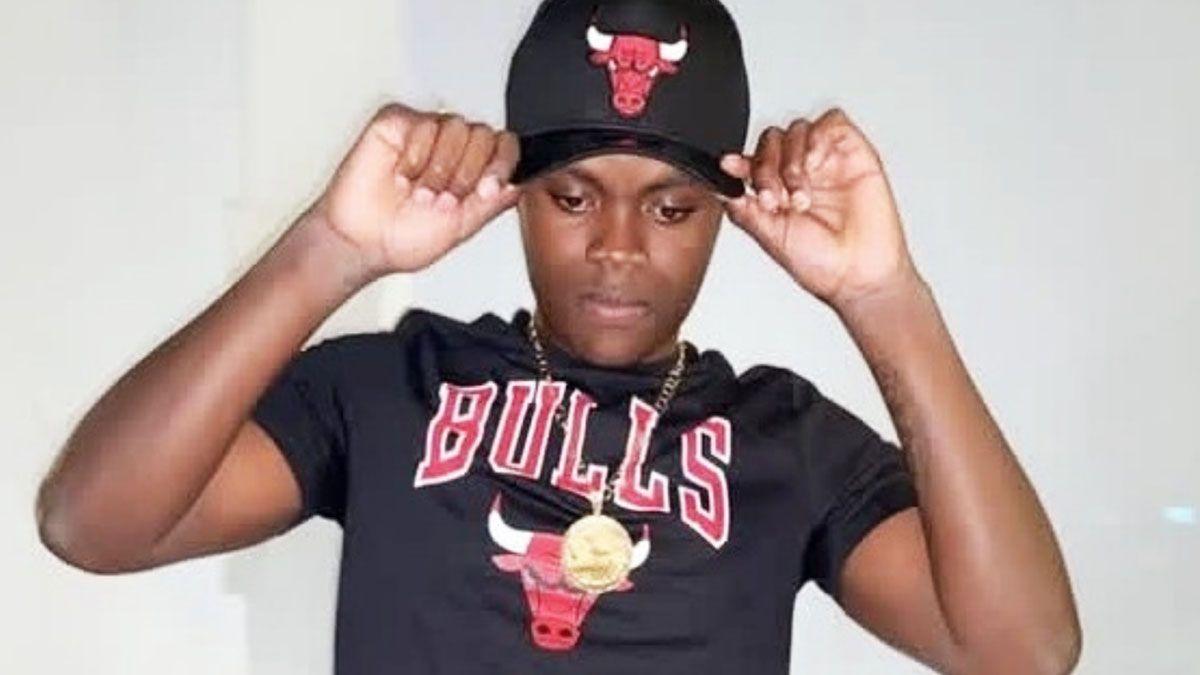 Khadeem Parris, a teenager, poses in front of the camera. He wears a Chicago Bulls-branded black T-shirt and a black baseball cap. The T-shirt is printed with the word "BULLS" in red lettering and the red face of a bull. The cap also has the red face of a bull printed on it. Khadeem holds the edges of the baseball cap with his fingertips. He wears a chain round his neck, and a white wall is behind him.