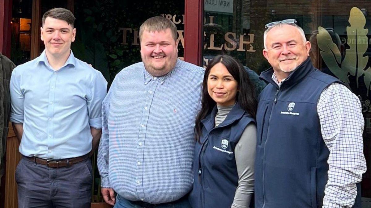 Jordan stood on the left with three colleagues. They are all dressed smartly in blue attire and stood outside The Welsh House restaurant where they work.