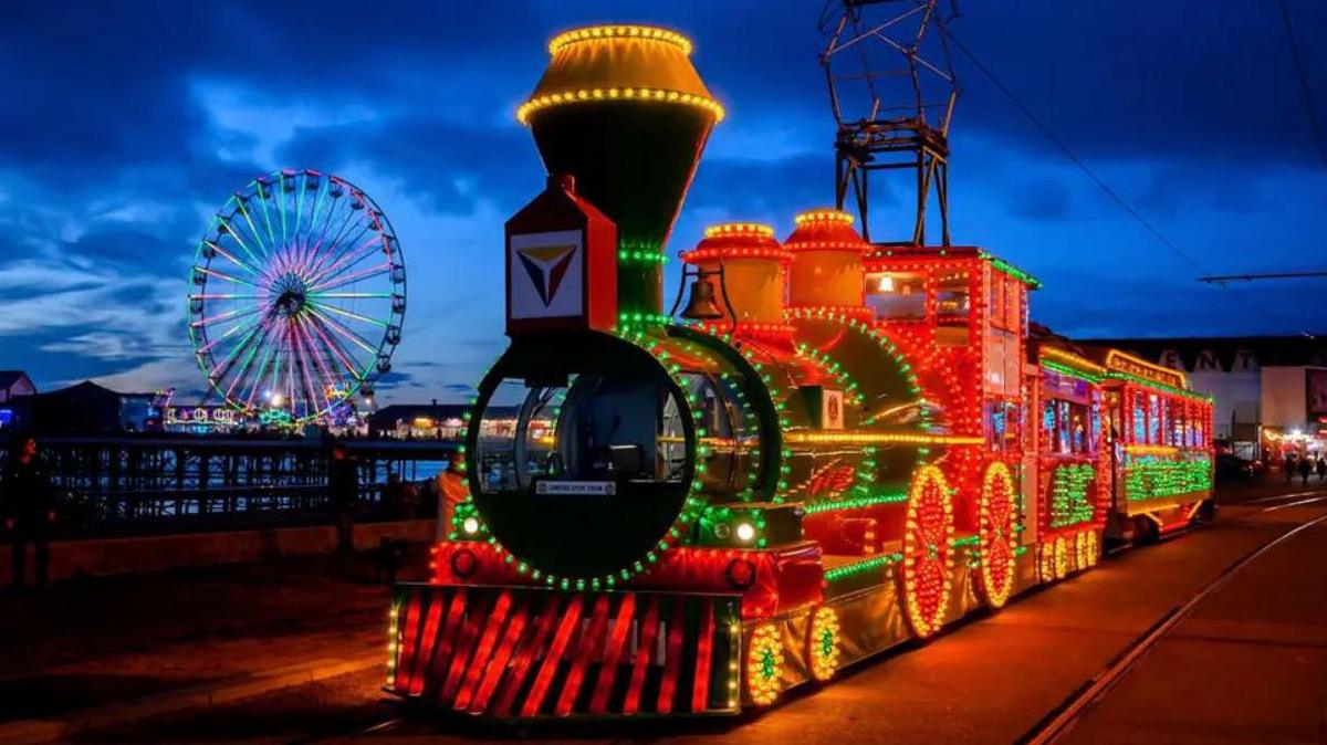 An illuminated train tram on Blackpool promenade with Central Pier's Big Wheel lit up in the background.