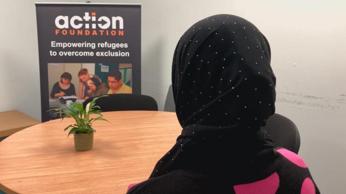 A woman the BBC has agreed to call Malali, facing away from the camera to protect her identity, wearing a black hijab and a pink and black spotty shirt. She is sitting at a desk with a small pot plant on it, with a banner for the charity Action Foundation ahead of her.