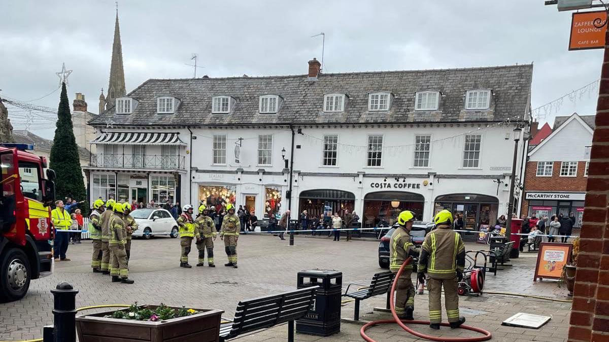 A large number of firefighters standing outside Market Place, Saffron Walden. You can see a white building, including Costa Coffee, park benches, a paved area, a Christmas tree to the left, Christmas decorations handing from buildings and a fire engine to the left. 