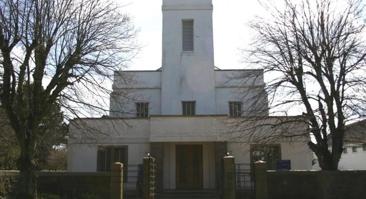 The large white church is seen in a pyramid shape with windows and a large-double door, framed by two large trees 