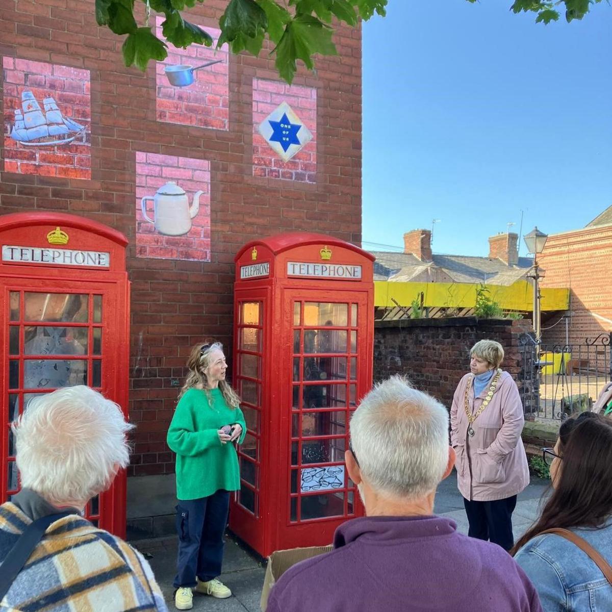 Artist Hannah Fox, in green top, standing between the phone boxes her art installations