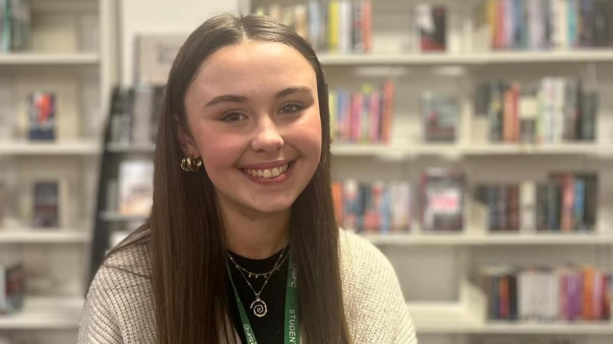 A head and shoulders image of Niamh, who is smiling into the camera. She is wearing a knitted cream-coloured cardigan over a black T-shirt. In the blurred background behind her are several bookshelves.