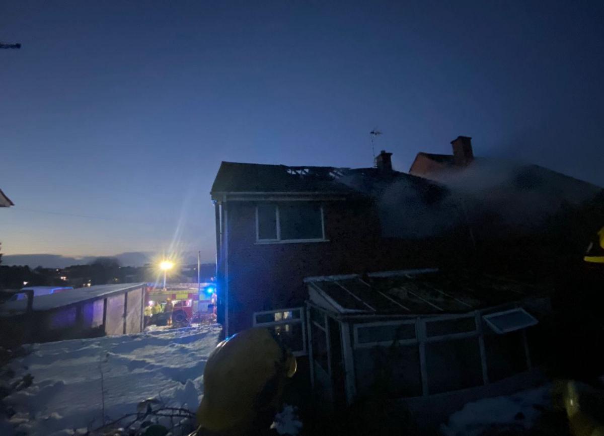 Firefighters in full gear look at a damaged house. It has a hole in the roof and a window blown out. Debris can be seen on top of the conservatory. A fire engine can be seen in the background with its blue lights on.