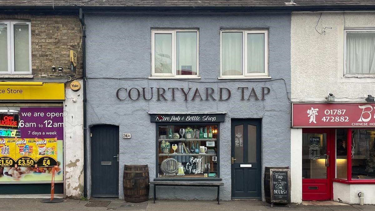 Courtyard Tap is an old grey building, and part of a row of shops. It has two blue/grey doors.