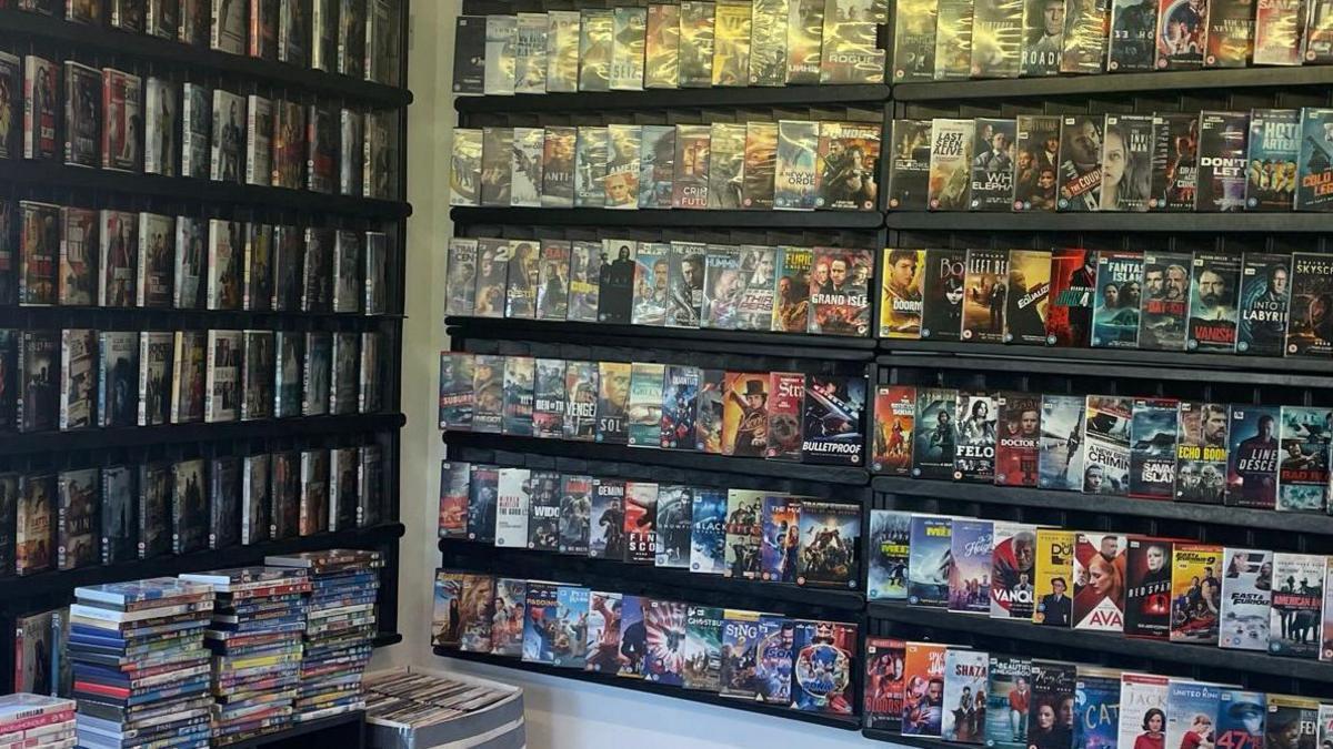 DVDs are piled high on top of shelves and on display in large black shelves on the wall.