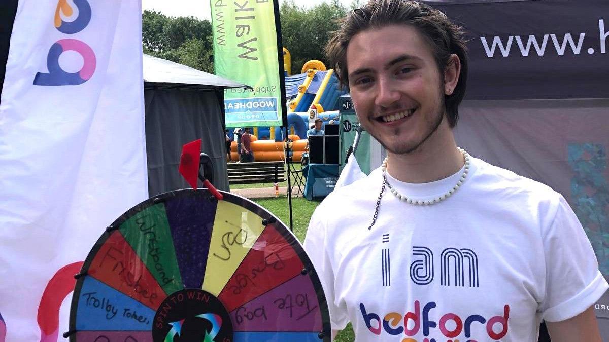 Russell Terrey, wearing a white T-shirt with "I am Bedford Giving" on it. He is standing by a wheel, in a park with rides behind him. He is smiling at the camera, and has short dark hair and a slight beard. 