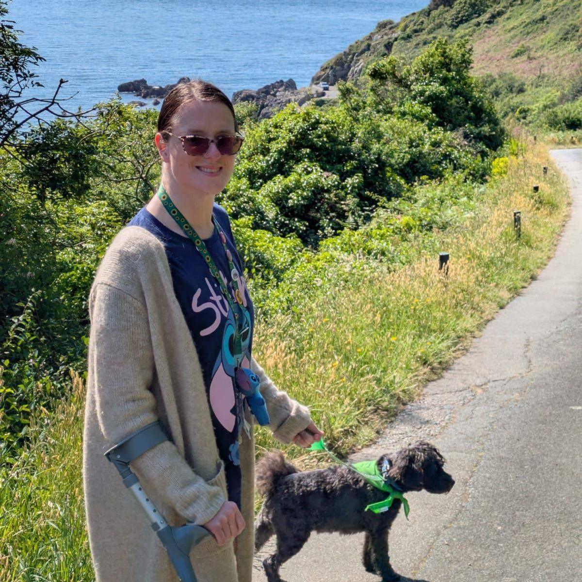A woman in light-sensitive glasses with a sunflower lanyard on and a crutch walks a small black dog down a country path, next to the coast of a small, rocky island.