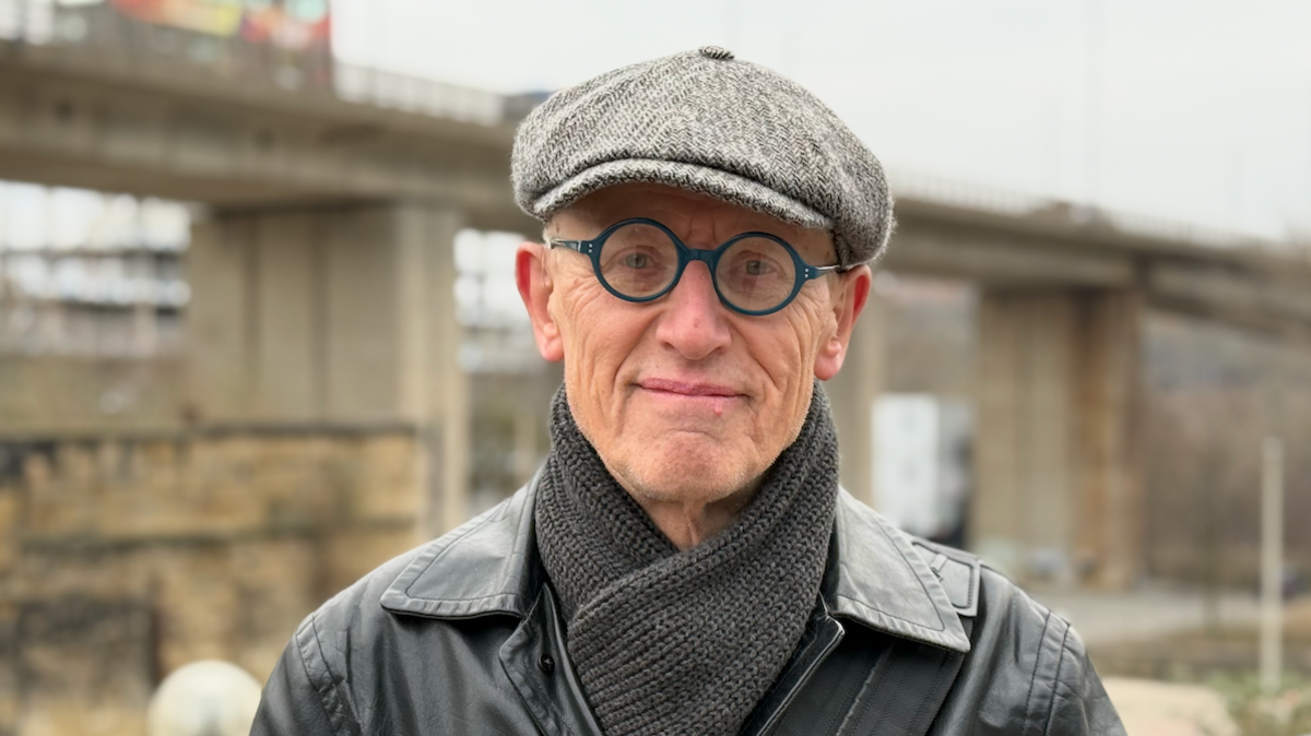 A man wearing a grey cap, a grey scarf and circular blue rimmed glasses stands in front of a beige-coloured concrete bridge.