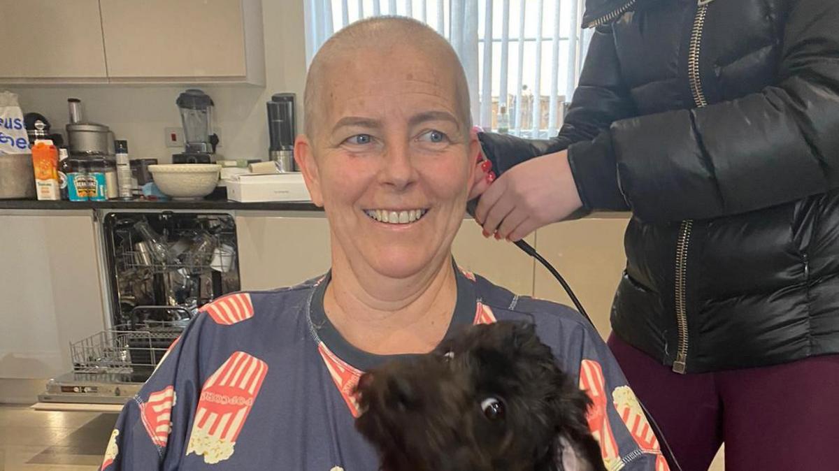 Lisa Noble smiling in a kitchen while having her head shaved during cancer treatment. She is wearing a blue T-shirt with illustrations of buckets of popcorn on it. A small black dog is sitting on her lap and has quickly turned to the camera with its eyes wide, creating an amusing scene. There is a woman with a black puffer jacket and maroon leggings holding an electric shaver next to her. Her face is off-camera.