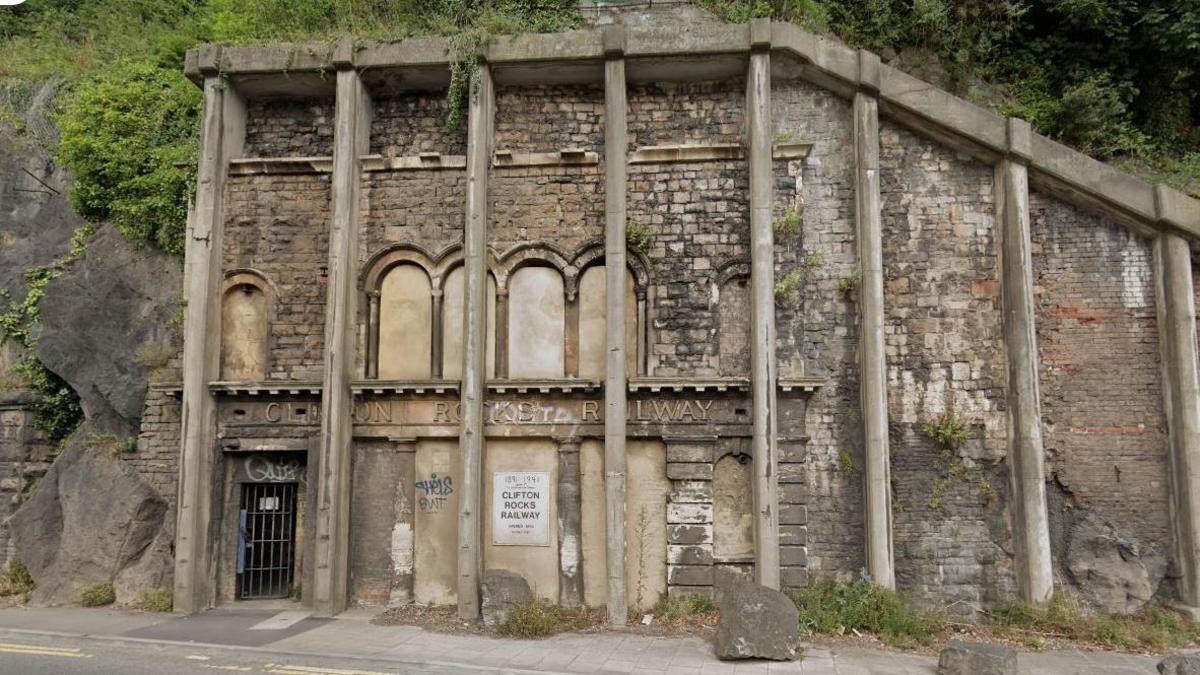 The remaining façade of the Clifton Rocks Railway. Its signage is barely visible underneath the concrete pillars installed to keep the structure from collapsing. Its arched tunnels and windows have been filled in and there is a rusty gate blocking the door.