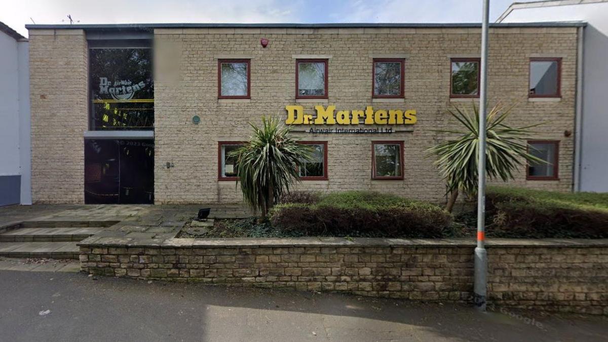 Light-brown brick-built two-storey factory with small square windows and yellow Dr Marten's logo on the front of the building. There are steps to the front entrance and there are two palm trees in the foreground.