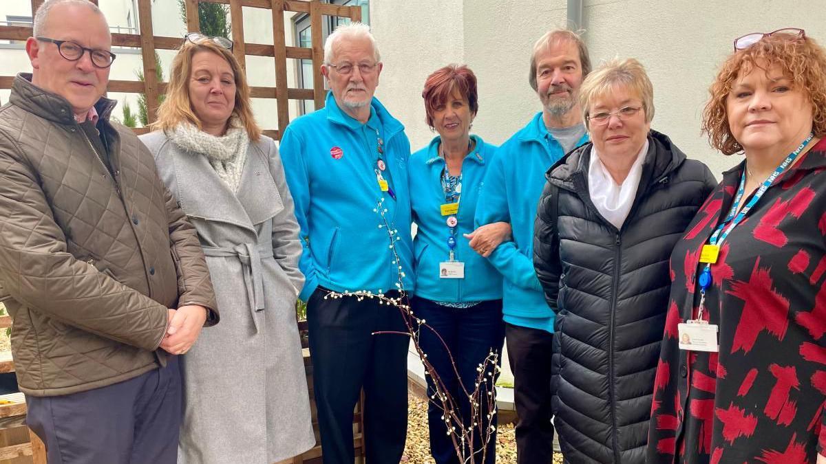 Five people stood around a potted acer tree. Three people in the centre are wearing volunteer uniforms. From left to right, the image shows Prof Steve Barnett, Clare Watts, volunteers Jeff, Tish and Jon, Jet Watts and Carol North. 